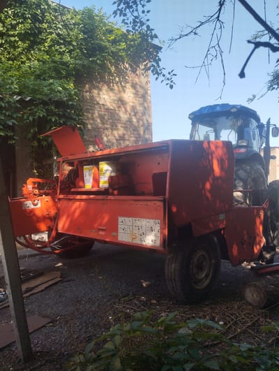 An orange chipper machine with its hopper open sits in front of a blue tractor under a bright sky. The chipper has two yellow containers inside it and several warning labels on its side. The tractor is large and appears to be a working vehicle. The scene is outdoors, with a brick wall covered in ivy in the background. The ground is rough and appears to be a mix of gravel and dirt. Sunlight casts shadows on the ground and the machinery.