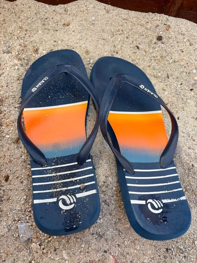 A pair of navy blue flip-flops with a gradient orange and blue design on the soles lies on a sandy beach. The flip-flops have white striped detailing and a white logo on the soles. The sand appears light brown and contains small pebbles and shells. The context of the location, Pallini Municipal Unit, Greece, is noted.