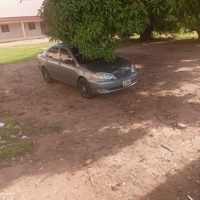 A gray sedan is parked on a dirt ground in front of a building in Wukari, Nigeria. The car is partially obscured by the branches of a large tree. The building appears to be a school or community center, with a light-colored facade and a red-tiled roof. There are a few scattered pieces of trash on the ground. The scene is well-lit by the sun, with shadows cast by the tree.