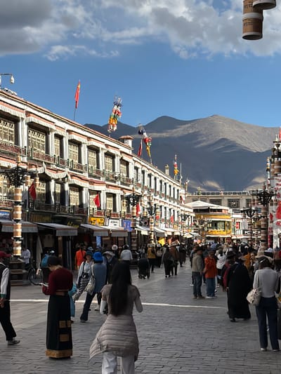 The image captures a bustling street scene in Chengguan District, China, characterized by traditional Tibetan architecture and a vibrant flow of people. The perspective looks down a paved street lined with multi-story buildings, backed by a large, barren mountain under a clear blue sky with scattered clouds.
The main subjects are numerous people walking along the street, many of whom appear to be engaged in a procession or pilgrimage. They are dressed in a mix of traditional and modern attire. Along the side of the street, attached to the buildings, are rows of cylindrical prayer wheels. Several individuals are seen actively turning these prayer wheels as they move, indicating a religious or cultural practice.
The buildings exhibit distinctive Tibetan architectural elements, with white facades, dark-framed windows, and decorative cornices. Colorful prayer flags, typical of Tibetan culture, are strung up in various locations, adding to the cultural atmosphere. A red flag, possibly featuring a single star, is also visible flying from a pole on one of the buildings.
Visible text includes characters on some of the prayer wheels. On one set, Chinese characters like "右" (yòu - right) and "上" (shàng - up) are discernible. On other prayer wheels, Tibetan script, including what appears to be "ཀ་" and "ཁ་", can be seen. Signs with illegible text are also present on the building facades.
The scene takes place on a bright, sunny day, likely in the late morning or early afternoon, given the clear illumination and the quality of the shadows. The majestic mountain range in the background firmly places the setting in a high-altitude, mountainous region, consistent with the Tibetan Plateau.
