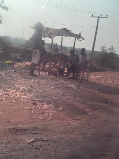 The image depicts an outdoor scene in Koton Karifi, Nigeria, characterized by an unpaved, reddish-brown dirt ground heavily scattered with wood shavings and chips. A bright, hazy sky is visible above.
In the mid-ground, a makeshift shelter composed of wooden poles supporting a light-colored, possibly torn fabric or tarpaulin provides shade. Beneath this shelter, a wooden structure resembling a table or workbench is present. Three individuals are positioned under the shelter; two are seated on small wooden stools, appearing to rest or engage in conversation. One seated individual wears a dark top and light-colored trousers, while the other wears a white top and dark trousers. A third individual is partially visible behind them.
To the left of the shelter, a tall, slender individual stands facing right, wearing a dark top, light shorts, and dark footwear. This person appears to be carrying a long, dark object, possibly timber, over their left shoulder.
In the background, an electricity pole with overhead power lines is visible, extending towards the right. Further distant, a larger metal lattice structure, possibly a transmission tower, can be seen against the sky. Sparse trees and vegetation line the horizon.