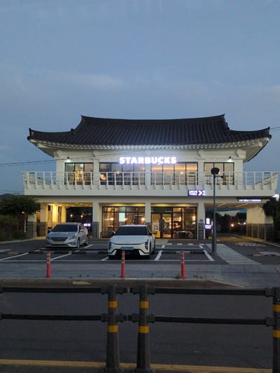 The image depicts a Starbucks store in Gyeongju-si, South Korea. The building is a two-story structure with a traditional Korean-style roof. The Starbucks logo is illuminated on the exterior. Cars are parked in a designated parking area. A street is visible next to the store, with a pedestrian crossing. The sky is visible in the background, suggesting it is dusk or dawn. A dark-colored metal guardrail is in the foreground, with orange reflective striping.