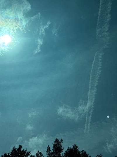 The image captures a sky scene over Longeville-lès-Saint-Avold, France, dominated by a deep cyan hue. In the upper-left quadrant, the sun appears as a brilliant, overexposed white light source, surrounded by a distinct lens flare and subtle light diffraction in shades of cyan and yellow. Scattered, wispy white cloud formations are visible throughout the sky, with some concentrated near the sun. A prominent, elongated white contrail extends vertically from the upper-right area downwards towards the mid-right, exhibiting a feathered, irregular texture. A small, faint, circular white object, consistent with the appearance of the moon, is discernible in the mid-right portion of the sky, below the contrail. The lower approximately 15% of the frame features the dark, silhouetted crowns of numerous conifer-like trees, forming an irregular, jagged horizon line. No human subjects, animals, or specific actions are depicted.