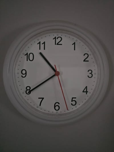 The image shows a close-up of a white analog clock mounted on a plain, gray wall. The clock has black Arabic numerals for hours 1 through 12 and small black dots marking the minutes. There are three hands: a black hour hand pointing between the 9 and the 10, a black minute hand pointing at the 10, and a thin red second hand pointing at the 6. The clock appears to be in a domestic or office setting. There is no indication of the specific location in Mäntsälä, Finland.