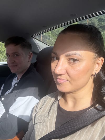 Two individuals are seated in the back of a car, with a sun-drenched outdoor scene visible through the rear window, suggesting they are likely traveling on a road in Beryozovsky, Russia. The person in the foreground, a woman with dark hair pulled back in a ponytail, is looking towards the camera. She is wearing a striped collared shirt over a dark t-shirt and a seatbelt. To her left, a man with short brown hair is also looking towards the camera, with a slight smile. He is wearing a black and white jacket over a white t-shirt, with a silver necklace visible. The interior of the car appears to be black, with sunlight creating strong shadows on the individuals and the car's interior. The background shows a dense forest with tall trees.
