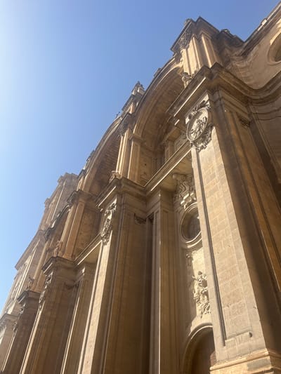 The image shows a low-angle shot of the facade of the Granada Cathedral in Granada, Spain. The sunlight casts strong shadows across the intricate stonework, highlighting architectural details such as columns, arches, and ornate sculptures. The sky above is a clear, bright blue. The composition emphasizes the scale and grandeur of the building.