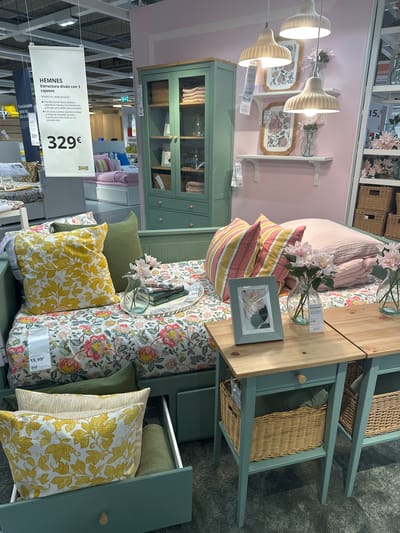 The image depicts a section of an IKEA store in Siero, Spain, showcasing a HEMNES daybed with three drawers. The daybed is dressed with a floral-patterned duvet cover and an assortment of pillows, including yellow patterned ones and green textured ones. A green duvet is neatly folded on the daybed, and a tray with books and a small vase of pink flowers sits on top of the bedding. To the right of the daybed are two light-colored wooden bedside tables with green bases, each featuring a drawer and a lower shelf with a woven basket. A framed picture of pink flowers is placed on one of the bedside tables. In the background, a tall, green glass-doored cabinet displays folded linens and vases. Hanging from the ceiling is a white IKEA sign advertising the HEMNES daybed, listing its price as 329€. The lighting suggests an indoor retail environment.