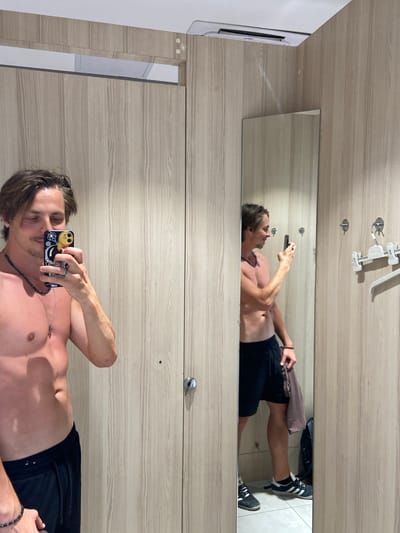 A shirtless man in black shorts lies on his back, holding a smartphone and taking a selfie in front of a mirror inside a locker room. The locker room has light-colored wood-grain lockers. The location is Bjeliši, Montenegro.