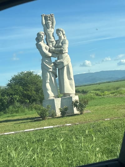 The image shows a stone monument, likely of socialist realism style, depicting three figures stacked atop one another. The monument is situated alongside a road or path in a rural or semi-rural location, possibly the city of Brezovo, Bulgaria, based on the user's query. In the background are fields of tall green grass and in the distance, some mountains. The sky is blue with a few scattered clouds. The photo appears to have been taken from inside a vehicle.