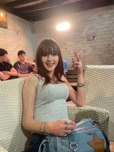 The image shows a young woman sitting in an armchair and smoking. She is wearing a grey tank top, jeans, and several rings and bracelets. She has long brown hair with bangs. In the background, two young men are sitting on a bench. The setting appears to be a dimly lit indoor space with white brick walls and a wooden ceiling. A sign with the words "RISE AND SHINE" and an image of a rooster is visible on the wall. There are no specific landmarks or features that definitively place this scene within the city of Hertsmere, United Kingdom.