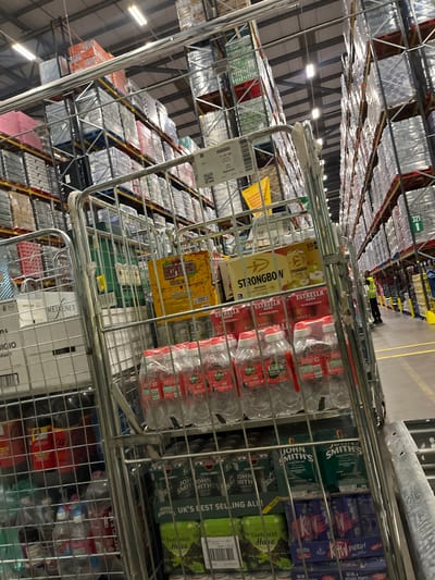 The image shows a busy warehouse environment, likely a distribution center, with tall shelves stacked high with various products. In the foreground, a metal cage trolley is partially filled with drinks, including bottles of Volvic water and cans of Estrella Damm beer. Other items visible in the trolley include boxes of Strongbow cider and John Smith's Tadcaster. In the background, more shelves laden with products are visible, wrapped in plastic. A person wearing a high-visibility vest is seen in the distance, suggesting ongoing operations. The overall impression is one of organized commercial activity, indicative of a logistics hub. The image was taken in the city of Northampton, United Kingdom.