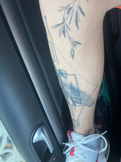 The image shows a person's lower leg with tattoos resting against the interior door panel of a vehicle. The door panel is black with a silver-colored accent strip and a silver door handle. A white athletic shoe with red accents is partially visible in the background. The tattoos on the leg include what appears to be an olive branch and a bird, possibly an eagle, within geometric shapes. The location is not evident from the image.