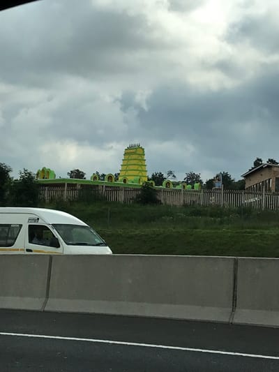 The image displays the Shree Radha Shyam Mandir, prominently situated in Sandton, Johannesburg, South Africa, visible from the adjacent N1 highway near the Rivonia Road interchange. The temple is characterized by its bright yellow and vivid green exterior, featuring a tall, multi-tiered central tower and smaller, dome-like structures along its roofline. It stands on an elevated grassy embankment, partially obscured by green foliage and a segmented wooden fence running along its perimeter. To the far right, a rustic-style building with a dark roof and light-colored walls is partially visible. In the foreground, a white minibus taxi with a horizontal yellow stripe is captured in motion, positioned behind a grey concrete crash barrier. A male driver is discernible within the taxi's cabin. The scene is observed from a paved road surface, with a white lane marking visible. The sky above is overcast with dense grey clouds.