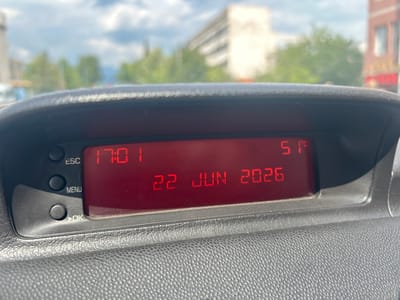 A close-up, eye-level shot captures the digital display of a vehicle's dashboard, showing the time as 17:01, the date as 22 JUN 2026, and the temperature as 51 degrees Celsius. The display is a bright red LED screen, situated within a dark grey, textured plastic housing. To the left of the display are several black, circular buttons labeled "ESC," "MENU," and "OK." The background of the image is blurred, indicating a focus on the dashboard. Visible through the car's windshield is an outdoor scene with a partly cloudy sky, trees, and buildings. The location is specified as Struma, Bulgaria.