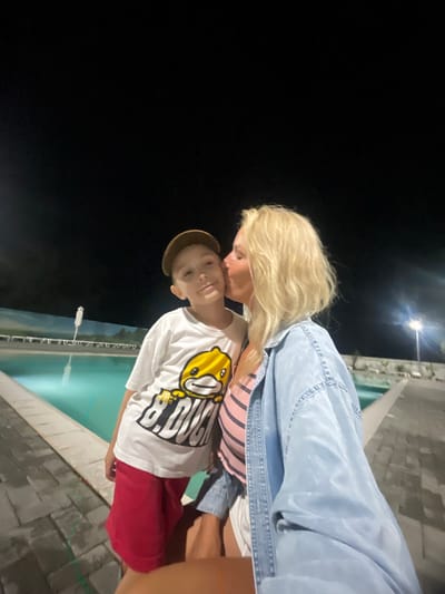 This image features two people, a woman and a child, in an outdoor setting at night. The woman, with blonde hair, is leaning down to kiss the child on the head. She is wearing a denim jacket over a striped shirt. The child, a boy, is wearing a white t-shirt with a cartoon character and black text, red shorts, and a tan baseball cap. They appear to be near a pool, indicated by the turquoise water and tiled edge visible in the background. The lighting suggests it is nighttime, with a bright light fixture visible in the distance. The scene evokes a sense of affection and a relaxed atmosphere. There is no visible text in the image.