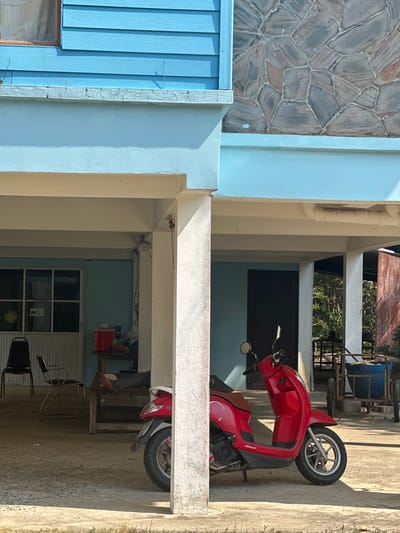 The image shows a red motorcycle parked in a covered outdoor area of a building in Ao Hin Khok, Thailand. The building has light blue walls and a textured stone facade. There are windows and doors visible in the background, along with chairs and other objects. The ground is concrete.