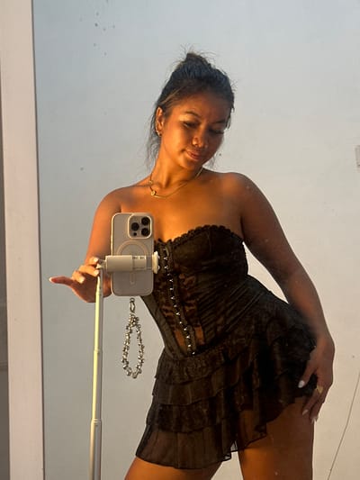 A woman is holding a smartphone attached to an extended selfie stick, likely taking a picture in a mirror. The woman is wearing a strapless black dress with a tiered, ruffled skirt. She is also wearing a gold necklace and a gold ring. Celuk, Indonesia is not discernible from the image.