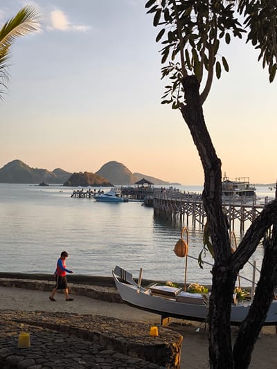 A person is walking on a paved pathway next to a body of water. The person is wearing a blue and red shirt and black shorts. In the background, there is a long wooden pier with people and boats. The water is calm and reflects the sky, which is a soft orange and yellow hue, indicating sunset. Islands with rolling hills are visible in the distance. The foreground features a light-colored boat on the sand, with some containers holding what appears to be food items covered with ice and banana leaves. There are also two woven baskets hanging from wooden poles. The scene is located on the coast of Labuan Bajo, Indonesia.