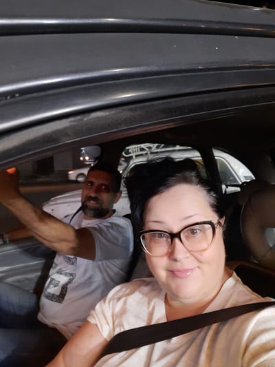 Two people are inside a vehicle. The person in the foreground is wearing glasses and a seatbelt. The person in the background appears to be exiting the vehicle. The background shows a street with other vehicles and lights, suggesting a nighttime setting. No information suggests the location is Cairo, Egypt.