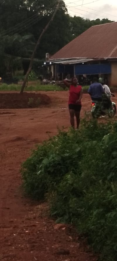 A medium shot shows a woman standing in the middle of a dirt road in Adikpo, Nigeria. She is wearing a red tank top and black shorts. Her right hand is on her head. Behind her, a man in a blue shirt and another person wearing a white shirt are on a motorcycle. The motorcycle is green and white. The background shows a building with a red roof and a blue awning. Trees and vegetation are visible. The sky is overcast.