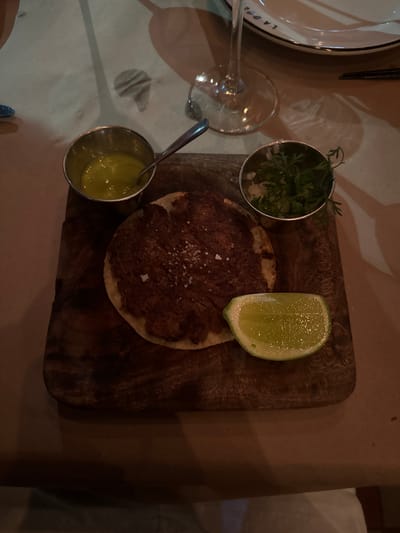 A top-down shot depicts a culinary scene, likely at a restaurant, though the presence of an individual's fingers at the bottom left suggests it could also be a home setting. The image features a wooden serving board, upon which a round dish, possibly a tostada, is placed. The dish is topped with a dark-colored spread, sprinkled with salt. Alongside the dish, there's a lime wedge and two small metal containers. One container holds what appears to be a green sauce, while the other contains fresh herbs, seemingly cilantro. The location is unspecified, but the details may indicate a dining establishment in Guadalajara, Mexico, based on the assumed dish presentation. Other elements present are a plate, cutlery, and a wine glass, placed on a neutral-toned surface or table.