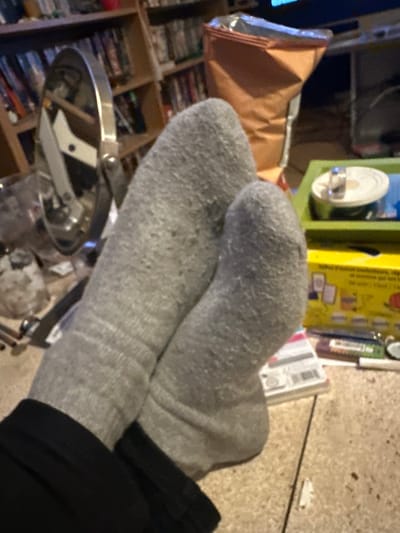 The image shows a pair of gray socks crossed over each other, with the cuffs visible, worn on a leg clad in black pants. In the background, there's a shelf filled with what appears to be DVDs. There is a brown bag of chips, and also a metal contraption. The floor is carpeted. There is no information to suggest that this image was taken in Gif-sur-Yvette, France.
