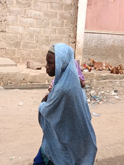 The image depicts an outdoor scene in Zango, Nigeria, under bright daylight. A young person stands in the foreground, facing left in profile. They wear a light blue head covering with white star patterns, over a visible grey and white striped cap. A purple garment is partially visible behind the primary subject. The ground is unpaved, composed of light brown dirt or sand, with scattered pieces of litter. To the left, a light-colored concrete block wall, marked faintly with "VCH-A-26," rises above a low concrete structure on which a loose concrete block rests. To the right, two light brown goats are positioned, resting on a raised concrete surface. Behind the goats, a building wall displays a reddish-pink upper section and a light grey textured lower section. Accumulations of refuse, including plastic and other debris, are present on the ground adjacent to the goats.