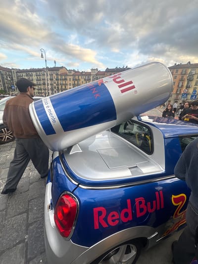 A person stands next to a blue and silver car with "Red Bull" prominently displayed on the side. A large replica of a Red Bull can is placed behind the car, appearing to touch the person's shoulder. The background features buildings and other individuals, suggesting an urban environment. The architecture, particularly the buildings, is consistent with the architectural style found in Turin, Italy. The scene likely shows a promotional event.