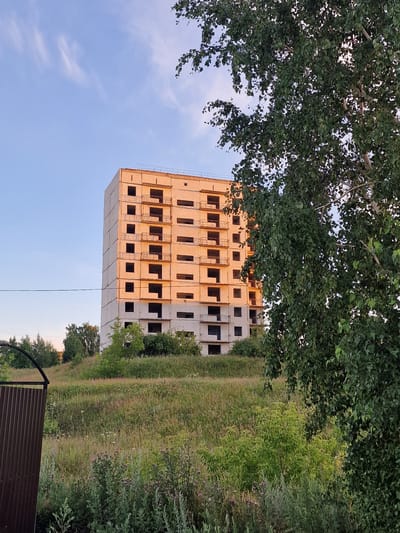 The image depicts an unfinished, multi-story concrete building under a clear sky with sparse, wispy clouds. The building, which appears to be residential, has numerous dark, empty window openings and exposed balconies. It is situated on a grassy hillside with scattered trees and vegetation. A tall, leafy tree is prominent in the foreground on the right side of the frame, partially obscuring the building. A metal gate, made of corrugated material, is visible on the left. The lighting suggests it might be late afternoon or early morning, with the side of the building illuminated by warm sunlight. There are no people visible in the image. The scene appears to be in a semi-rural or developing urban area. The location is identified as Kashgar City, China.