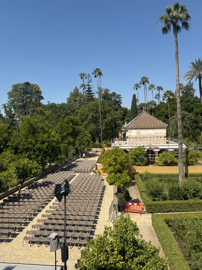 This is an outdoor scene in the gardens of the Alcázar of Seville, Spain. In the foreground, a large area is set up with rows of empty brown chairs, suggesting a performance or event space. A tall speaker on a stand is positioned in front of the chairs. Beyond the seating area, a pathway leads towards a building that appears to be undergoing renovations, with scaffolding around it. The building has a tiled roof and a pale facade. The background is filled with lush greenery, including numerous palm trees and other trees, under a clear blue sky. Several people are visible walking in the distance, and some are near the building. A orange construction barrier is placed on a path.