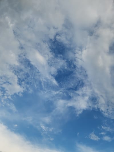 A photograph captures a wide expanse of the sky, predominantly featuring a mix of white and gray clouds against a vibrant blue backdrop. The clouds are varied in texture and density, with some appearing wispy and translucent, while others are more solid and billowy. The lighting suggests it is daytime, likely late morning or early afternoon, given the brightness of the blue sky and the way the sunlight interacts with the clouds. There are no discernible objects, people, or structures in the frame, making the sky the sole subject. The scene evokes a sense of openness and natural beauty. The weather appears to be partly cloudy, indicating a dynamic sky. No text is visible within the image.