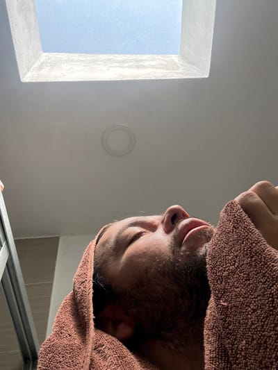 The image, taken in San Juan Cuautlancingo, Mexico, captures an upward-angled view of a person's head, primarily covered by a brown, textured towel. The person's face, visible from the chin to the forehead, shows stubble or a short beard. Their eyes are closed or cast downwards, and their lips are slightly parted, conveying a sense of relaxation or rest, likely after a shower or bath. A hand can be seen gripping the towel at the top of their head. The towel itself is a prominent feature, displaying its woven terry cloth texture.
The background consists of a light-colored ceiling or wall. To the left, a bright rectangular opening, likely a skylight or window, reveals a clear blue sky, indicating daytime. The frame of this opening is light-colored, possibly white, and appears to have a slightly textured or frosted finish. A circular recessed light fixture is mounted on the ceiling to the right of the skylight. In the bottom right corner, subtle hints of light brown or beige tiles and a metallic frame are visible, further suggesting an indoor setting, likely a bathroom. There is no visible text in the image.