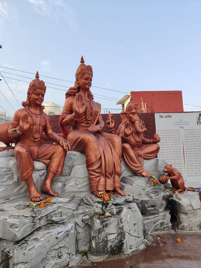 This image depicts a statue of Hindu deities, likely in Gurgaon, India. The central figure, a goddess with multiple arms, is seated. To her left, a male deity is seated, and to her right, Ganesha, the elephant-headed god, is also seated. In front of Ganesha, a small statue of a monkey is shown holding a round object. The statues are made of a bronzed material and are placed on a rocky, textured base. Several orange marigold flowers are scattered around the base of the statues, suggesting a religious offering. In the background, a red building and some power lines are visible, along with a clear blue sky. A large white banner with text in Hindi is partially visible behind Ganesha and the monkey statue. The overall scene suggests a public shrine or a religious monument.