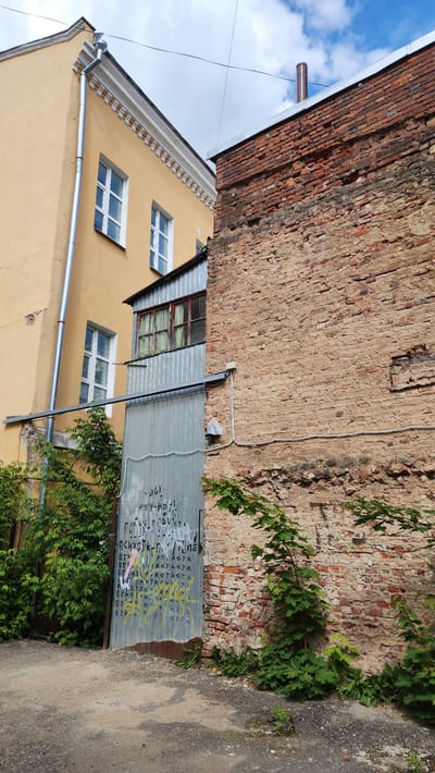 This image shows a partial view of two adjacent buildings in Smolensk, Russia. The building on the left is a two-story structure painted in a light yellow hue. It has white-framed windows. A silver gutter runs vertically down the side of the building. To the right is an older brick building with visible weathering and a faded color. A section of the brick wall has plant growth. A corrugated metal door or panel is leaning against the brick wall, covered in graffiti. The ground to the right is paved. A small patch of overgrown plants is in front of the metal panel. The sky with clouds is visible above the left building.