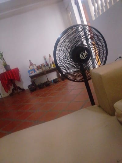 This image depicts an indoor scene with a floor tiled in terracotta. In the foreground, a portion of a light-colored sofa is visible. A black oscillating fan stands to the right of the sofa, with its grill and "LM" logo clearly discernible. To the left of the fan, a small table draped with a red cloth is set against a white wall. On the table, a potted plant can be seen. Further to the left, a long, dark wooden table holds various decorative items, including bottles, frames, and what appear to be small statues. The overall impression is that of a residential interior, possibly a living room or dining area. There is no specific information in the image to confirm the location as Atamo Sur, Venezuela.