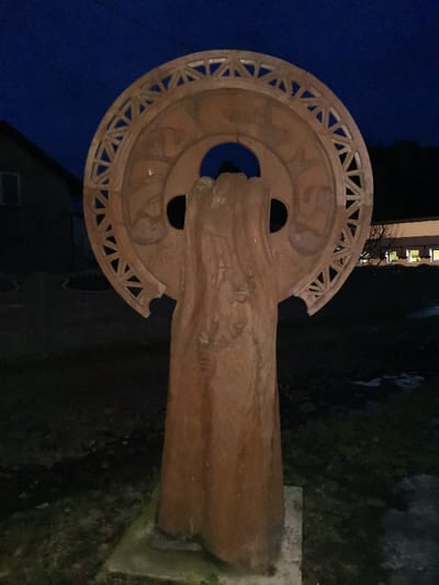 The image displays a large, weathered wooden sculpture positioned outdoors at night or dusk in Masty, Belarus. The sculpture features a tall, vertical pillar-like form supporting a prominent circular element at its apex. The circular section includes an intricately carved outer rim with repeating triangular openwork, and its inner surface contains relief carvings, two dark circular cutouts, and a central, irregular aperture. The main vertical shaft of the sculpture exhibits a textured surface, suggesting flowing drapery or abstract forms, with faint relief details visible. It stands on a light-colored, square base. In the left background, a dark building with a single rectangular window is partially visible. To the right, further back, a lighter-colored building with several illuminated rectangular windows is situated behind dark trees or shrubs. The ground surrounding the sculpture consists of dark earth or sparse grass, with some lighter patches. No individuals are present in the scene.