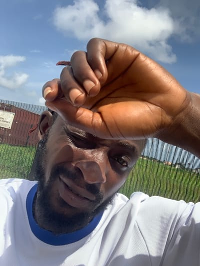 This is a close-up, selfie-style photo of a Black man outdoors on a sunny day. The camera is angled upwards, showing the man's face, a portion of his hand held up to his forehead, and his white t-shirt with a blue trim. He has a beard and is looking towards the camera with a slight smile. His hand, with dark skin and neatly trimmed nails, is blocking some of his forehead.
In the background, a black chain-link fence runs horizontally across the image. Beyond the fence, a green field is visible, suggesting an outdoor recreational area or sports field. The sky above is bright blue with scattered white clouds, indicating fair weather and likely daytime. There's a hint of buildings with red roofs visible behind the fence in the lower portion of the image, near a sign. The overall impression is one of a person enjoying a sunny day outdoors, possibly after some physical activity given the sweat on his forehead and the athletic wear. The location context is Sangotedo, Nigeria.