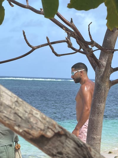 A man with a shaved head and sunglasses is standing on a beach. He is wearing pink shorts. The water is turquoise and there are trees and branches in the foreground. The location is potentially Santa Monica, Philippines, based on the image's style, though confirmation requires additional data. The photograph is taken from an unusual angle, rotated 90 degrees counter-clockwise.