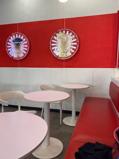 The interior of a restaurant in Kecamatan Piyungan, Indonesia, located at latitude -7.837 and longitude 110.474, features red and white walls with circular advertisements depicting beverages. There are four white chairs and three round, light pink tables. Two chairs are situated at the tables, while one chair is empty. A long red bench with a black sweater lying on top of it is adjacent to a white wall. The wall has a vertical crack in it. The scene is lit by artificial lighting.