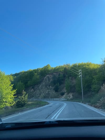 A roadside scene in Echinos, Greece, shot from inside a vehicle. A winding asphalt road with double white lines is visible on the right side of the frame. Adjacent to the road is a rocky cliff covered with dense green trees and vegetation. A wooden utility pole with multiple wires extends towards the left, against a clear blue sky. The photograph appears to be taken during daylight.