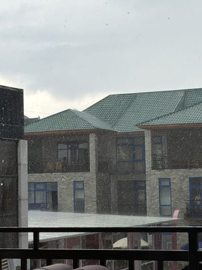 The image depicts a multi-story building with a green, tiled roof and stone facade, viewed through what appears to be a rain or snow shower. The building has multiple windows, some with blue frames, and balconies. A dark structure is partially visible on the left side of the image. In the foreground, there is a dark railing. The overall environment suggests an outdoor setting in a city, potentially Bosteri, Kyrgyzstan, given the architectural style and the weather conditions. The presence of what looks like snow or heavy rain falling obscures some details of the scene.