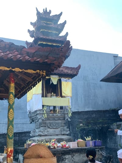 This image depicts a religious ceremony taking place outdoors, likely at a temple or shrine, in Sukasada, Indonesia. The central focus is a multi-tiered Balinese structure, possibly a meru or altar, adorned with intricate carvings and decorative elements. The structure is draped with white and yellow fabric. At its base, several woven baskets filled with offerings, including flowers and other symbolic items, are arranged on a stone platform. In the foreground, a large, domed woven basket is prominent. To the right, a man dressed in white traditional attire, holding incense sticks that are producing smoke, stands facing the altar. Partially visible at the bottom right is another person also in white. The background consists of a textured, light blue wall. The overall scene suggests a ritualistic offering or prayer in a tropical environment, given the sunlight and greenery visible.