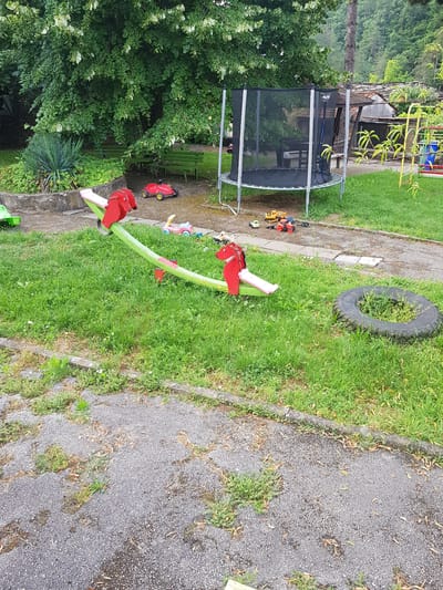 In Lomets, Bulgaria, a vibrant, slightly worn outdoor playground features a green and pink seesaw with red horse-shaped handles positioned on a lush, green grassy area. Scattered around the playground are various toys: a red toy car, a pink and blue toy vehicle, and several small construction vehicles, including a yellow excavator and a red truck. A large black trampoline with a safety net stands in the background, near a rustic wooden structure with a tiled roof. The environment is characterized by dense green trees providing shade, and the ground is a mix of gravel paths and grass. A discarded tire, partially overgrown with weeds, lies to the right of the seesaw. The scene captures a moment of playful stillness in a natural, slightly unkempt, outdoor setting.