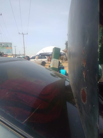 The image depicts an outdoor daytime street scene in Mista Ali, Nigeria, viewed from the interior of a vehicle. The immediate foreground shows the dark, cracked rear window of the observer's vehicle. To the right, a large, dark, textured object with a curved surface, chipped paint, and multiple circular perforations is partially visible.
In the midground, a bustling street environment unfolds. Overhead, numerous power lines stretch across the clear sky, supported by wooden utility poles. On the left, a large advertising billboard with text and images is visible. Below, several vehicles are present on what appears to be an unpaved or dirt roadside. A prominent white van or SUV is parked beneath a large, white market umbrella bearing the "NIVEA" brand logo in blue. Near this umbrella and vehicle, a person wearing a bright blue shirt is seated on the ground or a low stool, positioned adjacent to a distinct green and white striped pole. Other indistinct figures are scattered throughout the market area, suggesting commercial activity. The background features distant structures and minimal vegetation under a clear sky.