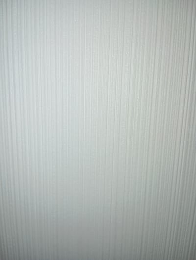The image displays a close-up, full-frame view of a white, textured surface. This surface features a pattern of vertical, parallel ridges and grooves. The texture appears somewhat rough or matte, with subtle variations in light and shadow highlighting the three-dimensional quality of the ridges. The overall impression is that of a neutral, man-made material, possibly wallpaper or a similar wall covering. There are no people, objects, or discernible environmental elements visible in the frame, making it a purely textural study. The location in Hlynytsi, Ukraine, cannot be determined from the visual information presented in the image.
