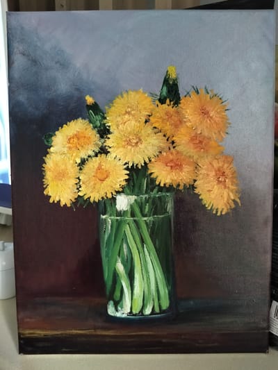 A close-up, eye-level shot captures an oil painting of dandelions in a glass vase. The dandelions, rendered in vibrant yellow and orange hues with textured brushstrokes, dominate the canvas. They are arranged in a clear glass vase filled with green stems. The background is divided, with the upper portion featuring a dark, moody gradient from gray to deep blue, and the lower portion a rich, dark brown. The scene is set on a wooden surface, indicated by the brown tones in the lower part of the painting, which suggests a still life composition. The painting appears to be on stretched canvas, with the texture of the fabric visible. There are no people present in the image. The lighting within the painting is soft, creating subtle highlights and shadows on the flowers and vase. The overall impression is one of rustic simplicity and natural beauty. There is no information provided about the location in Syktyvkar, Russia.
