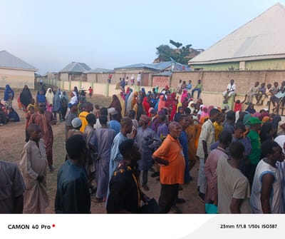 The image, likely taken in Wase, Nigeria, depicts a dense crowd of people gathered outdoors. A mix of adults and children are present, with many wearing traditional clothing. A large number of people are sitting atop a concrete wall behind the crowd. Buildings with corrugated metal roofs are visible in the background. The sky is overcast and light.