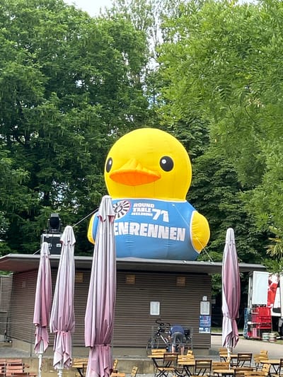 A large, inflatable yellow duck wearing a blue vest with "ROUND TABLE 71 HEILBRONN" and "RENNEN" printed on it is positioned above a small, wooden building with a red awning. The building has several closed windows and doors, and three large, closed pink and white umbrellas are standing in front of it. There are tables and chairs set up outside the building. In the background, there are trees and a white truck with red crates. A person is visible in the lower left corner of the image, bent over a bicycle. The scene appears to be outdoors, possibly at an event or fair in Heilbronn, Germany.