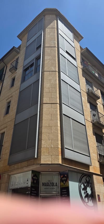 The image shows a multi-story building with a corner facade made of light brown stone blocks and grey metallic paneling. The building has multiple windows with closed grey roller shutters. On the ground floor, there are shopfronts with large glass windows. One of the shopfronts displays signage that reads "CHEZ MADZOLA PIRINEO GOURMET" and lists various products. The street or sidewalk is visible in the foreground, partially obscured by a blurry pink object, possibly a finger. The sky is clear and blue. The architectural style suggests an urban setting, likely in a European city. Based on the signage, this building is located in Huesca, Spain, at the "Chez Madzola Pirineo Gourmet" shop.