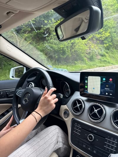 A close-up photograph taken from within a Mercedes-Benz car reveals a woman driving. The car's infotainment system displays a map with a location near Dobrini in the vicinity of Pastra, Bulgaria. The driver has her hands on the steering wheel, wearing a bracelet on her left wrist. The car's interior is beige, with a silver-colored dashboard. The view through the windshield shows a green, tree-filled outdoor environment. The rear-view mirror is visible in the upper-left corner of the frame.